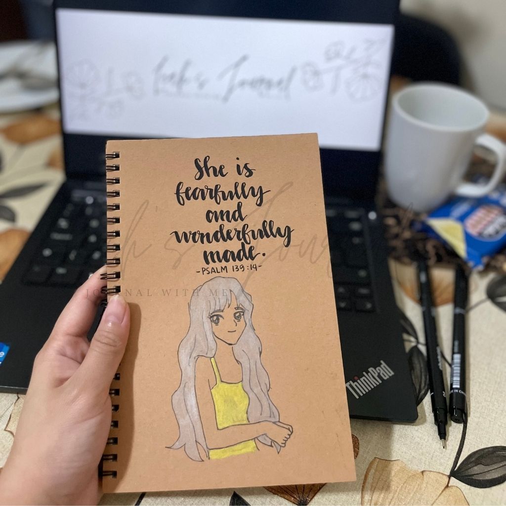 Notebook with illustration and text held by a hand, with a laptop and coffee cup in the background.