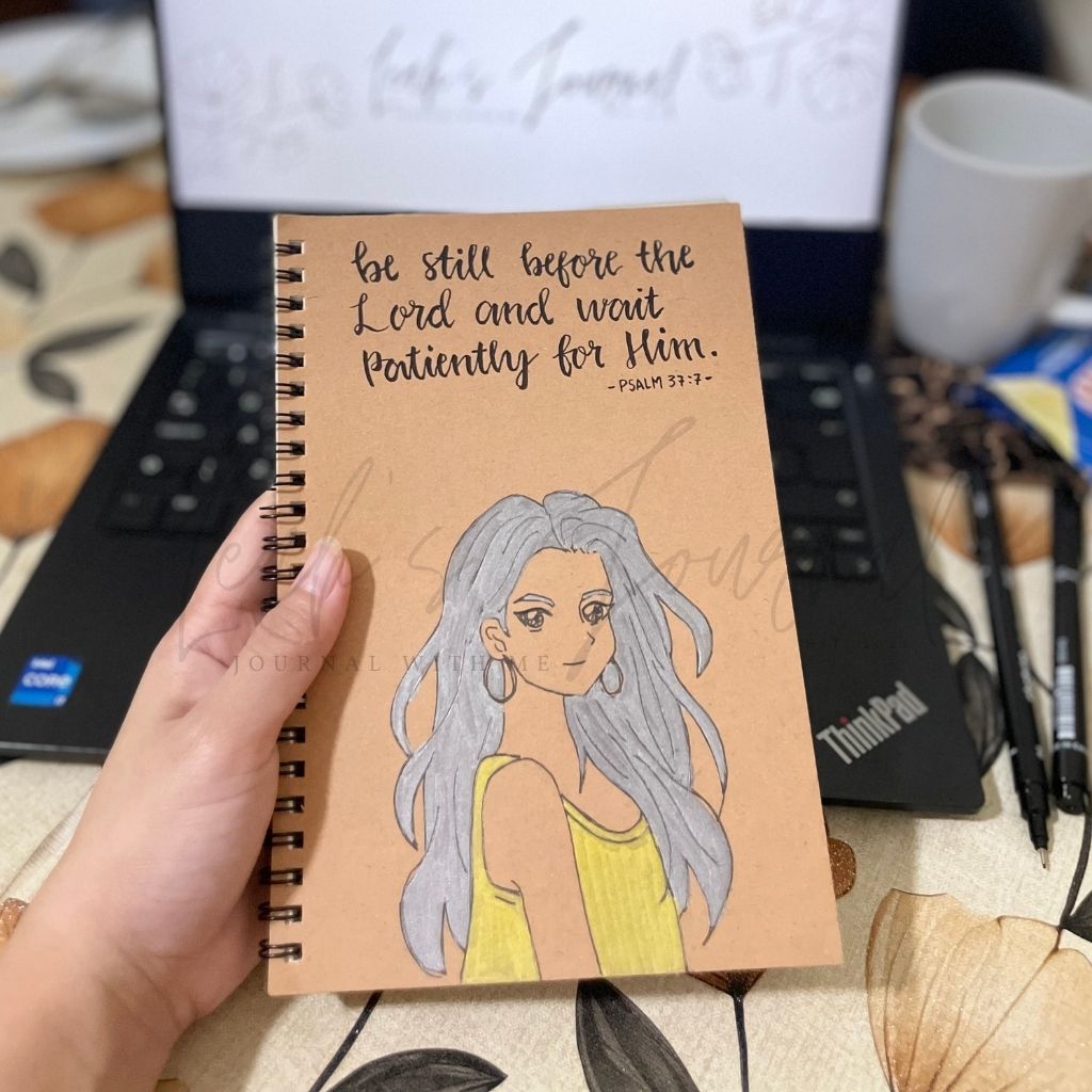 Notebook with illustration and text held by a hand, with a laptop and coffee cup in the background.