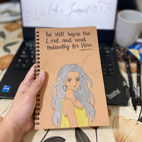 Notebook with illustration and text held by a hand, with a laptop and coffee cup in the background.