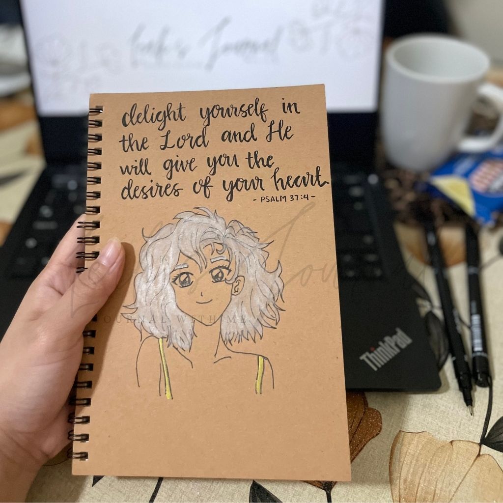 Notebook with a illustration and text held by a hand, with a laptop and coffee cup in the background.