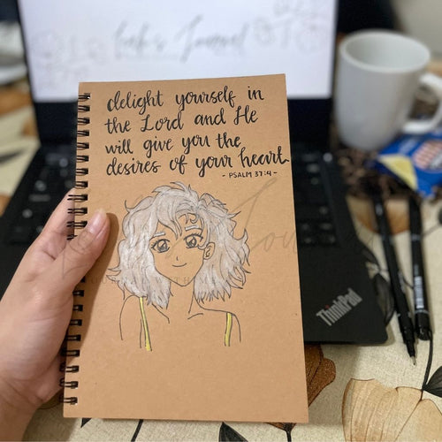 Notebook with a illustration and text held by a hand, with a laptop and coffee cup in the background.