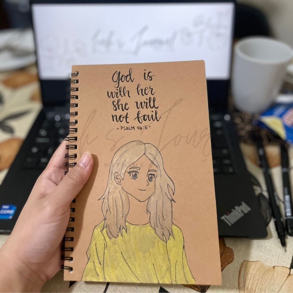Notebook with a drawing of a person and text 'God is with her, she will not fail' held by a hand.
