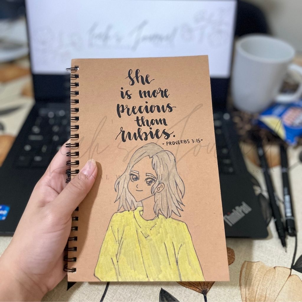 Notebook with illustration and text held by a hand, with a laptop and coffee cup in the background.