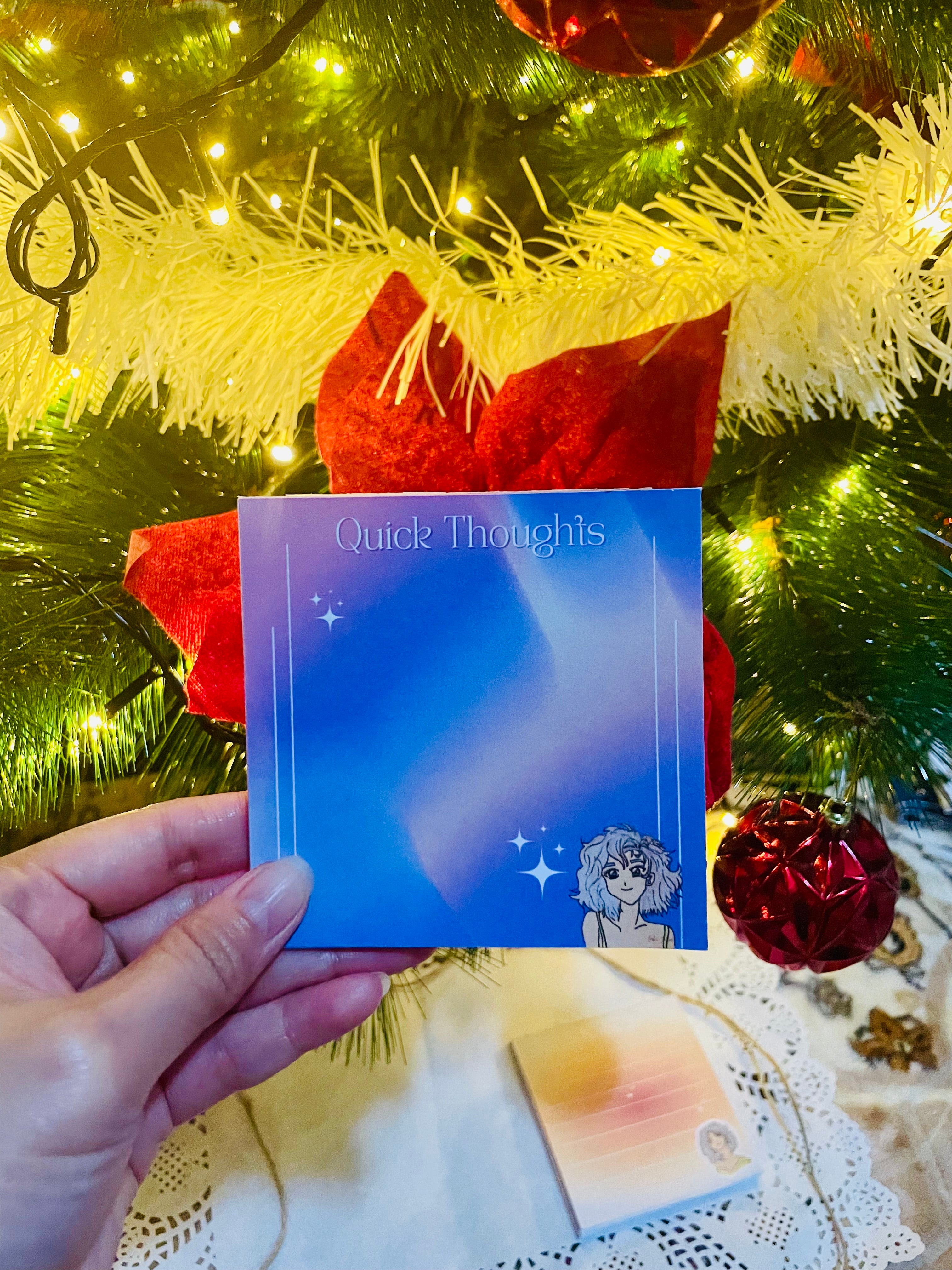 Hand holding a blue card with 'Quick Thoughts' text in front of a decorated Christmas tree.
