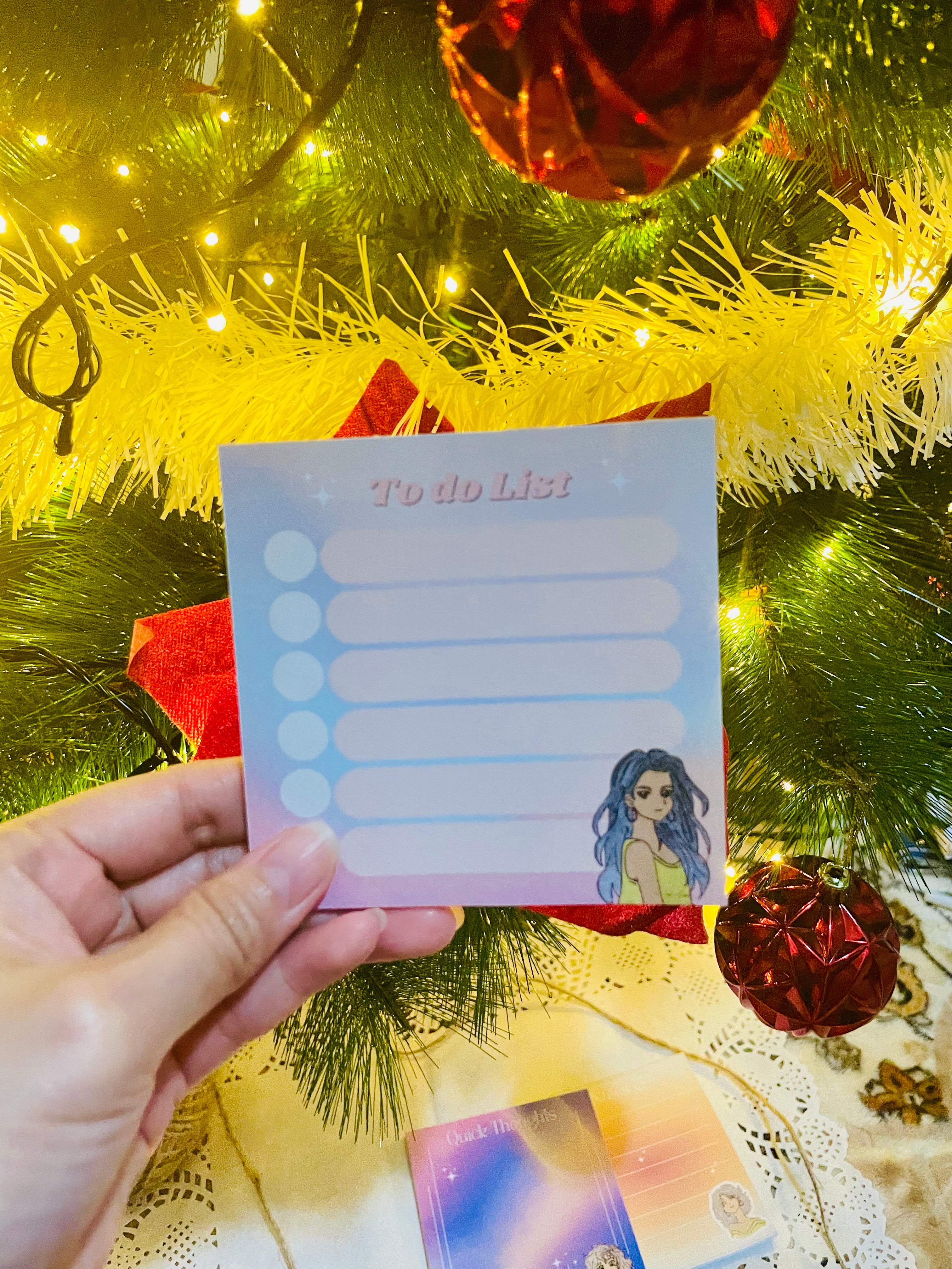 Hand holding a to-do list card with a character illustration in front of a decorated Christmas tree.