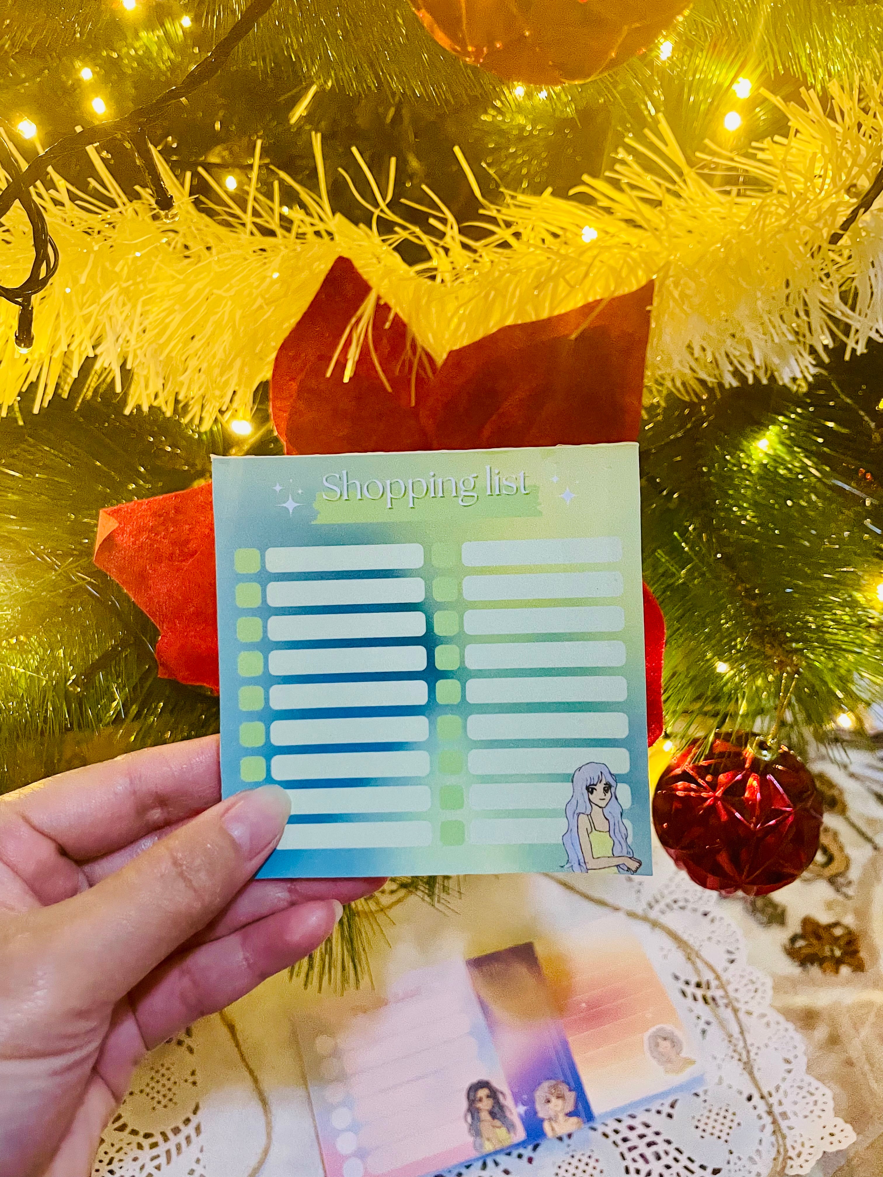 Hand holding a shopping list card in front of a decorated Christmas tree.