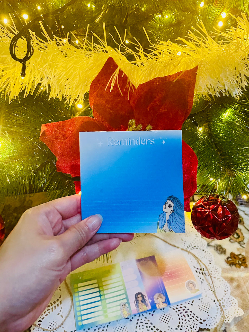 Hand holding a blue card with a character illustration in front of a decorated Christmas tree.