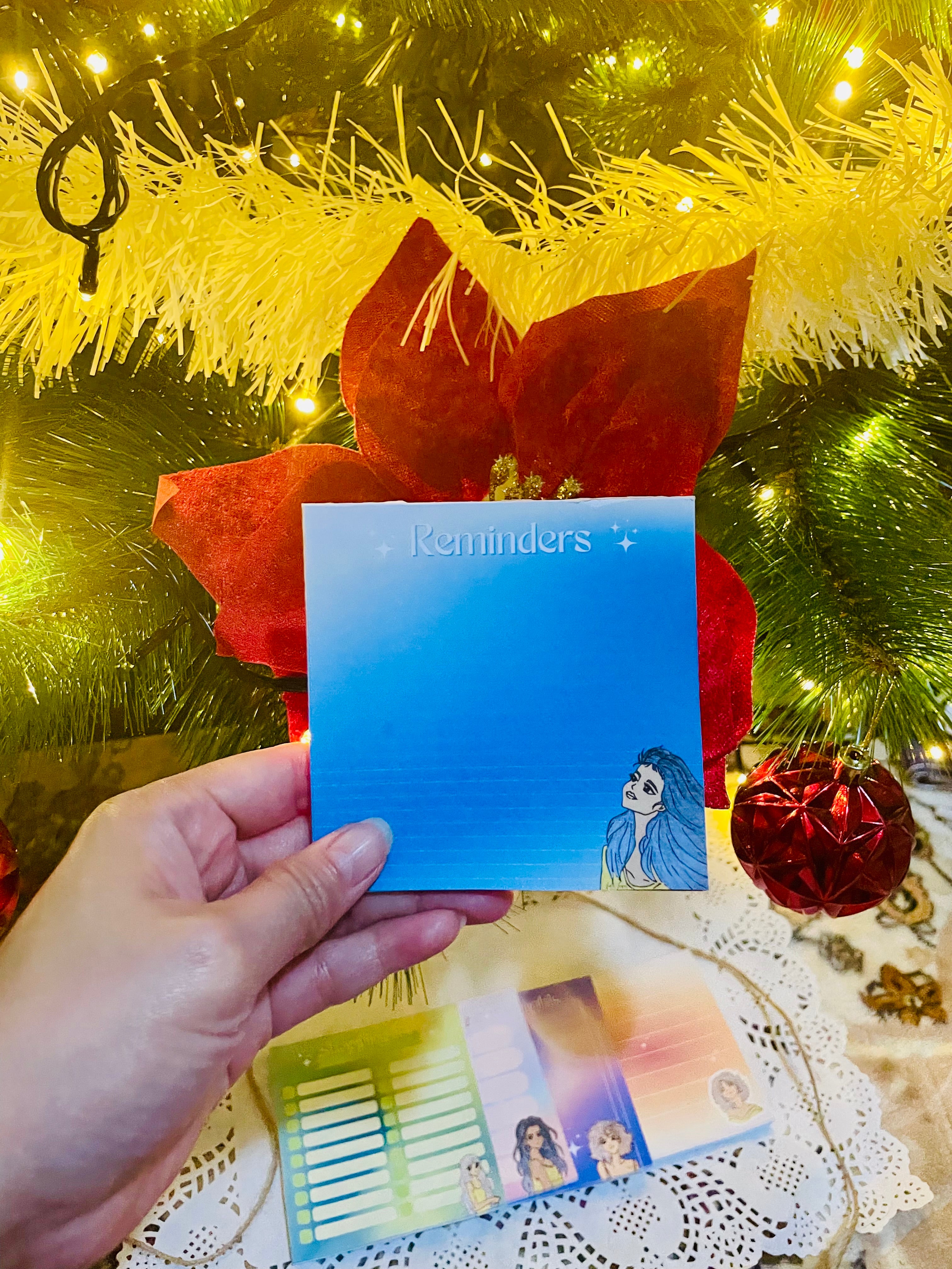 Hand holding a blue card with a character illustration in front of a decorated Christmas tree.